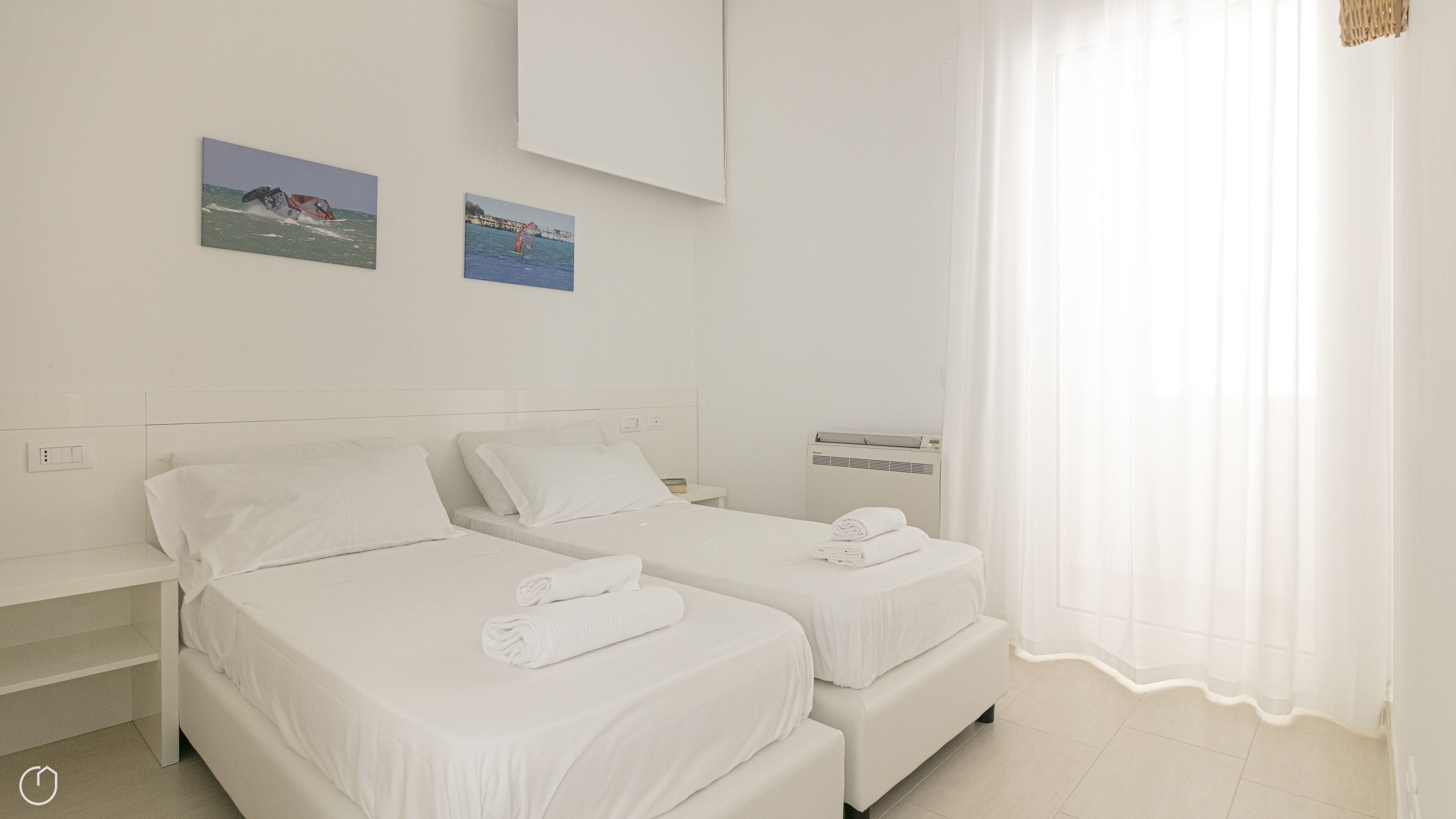 A hotel room with two single beds, a small bedside table, a wall-mounted air conditioner, and two framed pictures of coastal scenes.