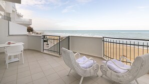 A balcony with a view of the beach, white wicker chairs, a small table with a book and a pink mug, and a white chair.