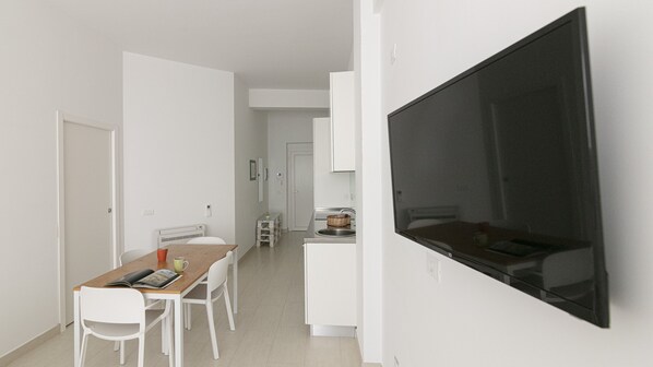 A modern, minimalist kitchen with white cabinetry, a large flat-screen TV mounted on the wall, and a dining area with a wooden table and white chairs.