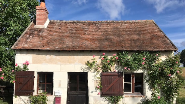 Une maison de campagne en pierre, avec un toit en tuiles, deux portes en bois et un petit jardin agrémenté de fleurs roses.