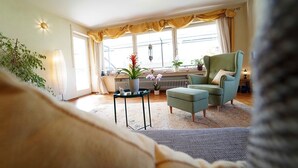 Una sala de estar acogedora con un sofá, un sillón verde, una mesita con plantas y una ventana con cortinas.