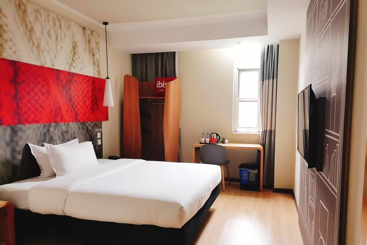 A hotel room with a bed, a desk with a chair, a wardrobe, a television, and a window with curtains.