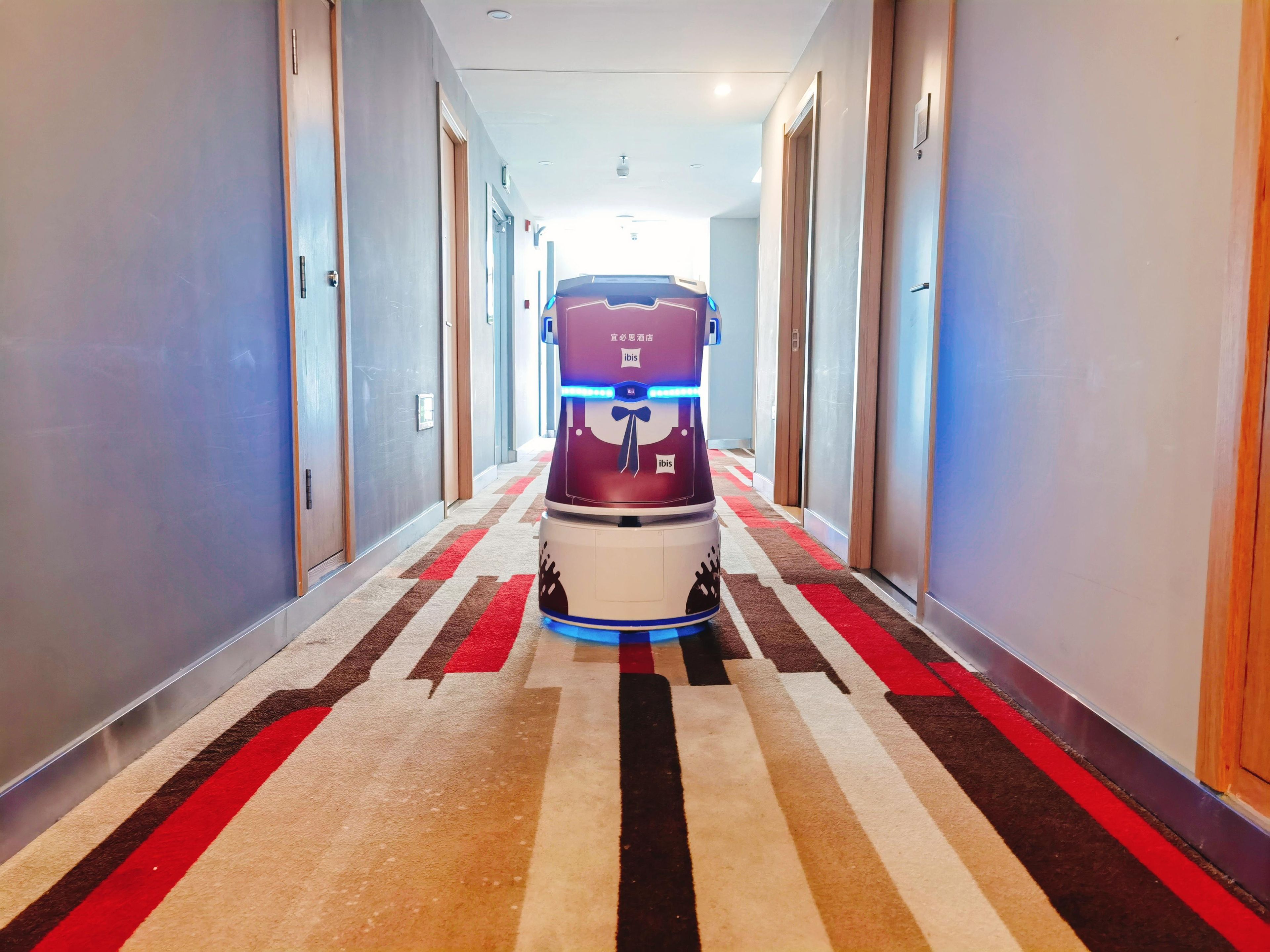 A hotel hallway with a robotic vacuum cleaner, doors on both sides, and a patterned carpet.