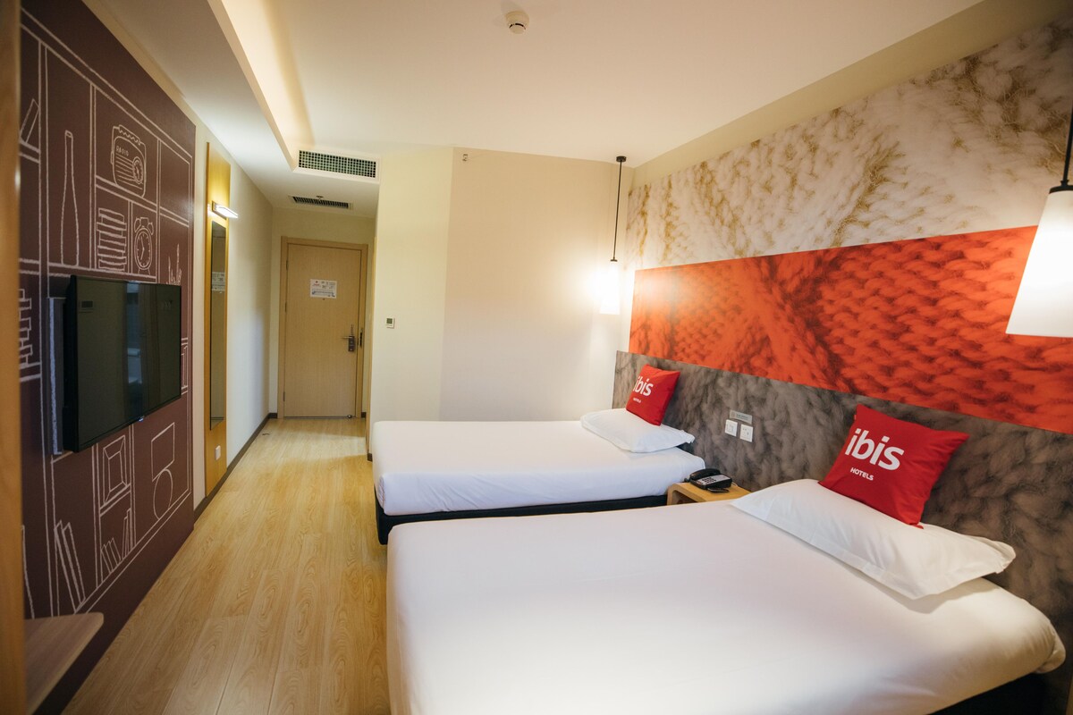 Hotellihuone, jossa on kaksi sänkyä, televisio, kuvioitu seinä ja punainen koristetyyny, jossa on hotellin nimi.