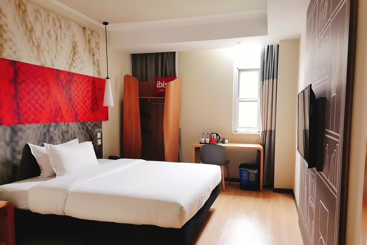 A hotel room with a bed, a desk with a chair, a wardrobe, a television, and a window with curtains.