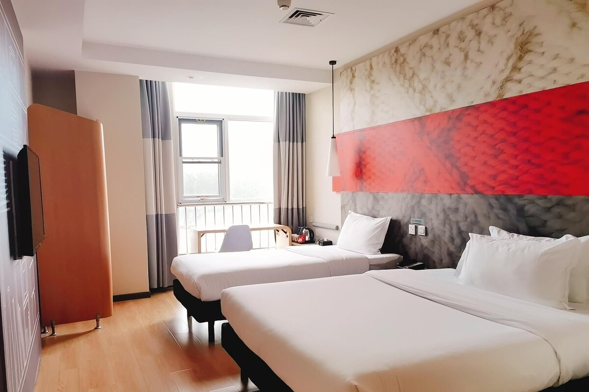 A hotel room with two beds, a wooden floor, a window with curtains, and a marble accent wall.
