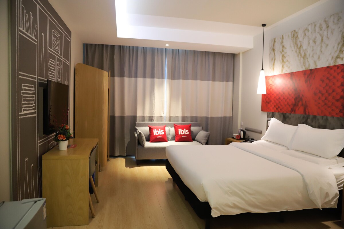 Quarto de hotel com cama, secretária, guarda-roupa, televisão e janela com cortinas.