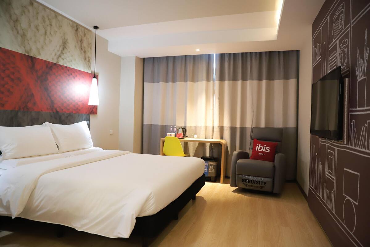 A hotel room with a bed, a desk, a chair, a TV, and a wall with a mural.