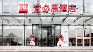 A hotel entrance with a glass facade, a red sign, and two flower arrangements.
