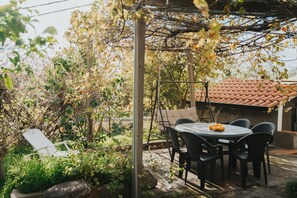 Un patio con mesa y sillas, rodeado de follaje y una pérgola.