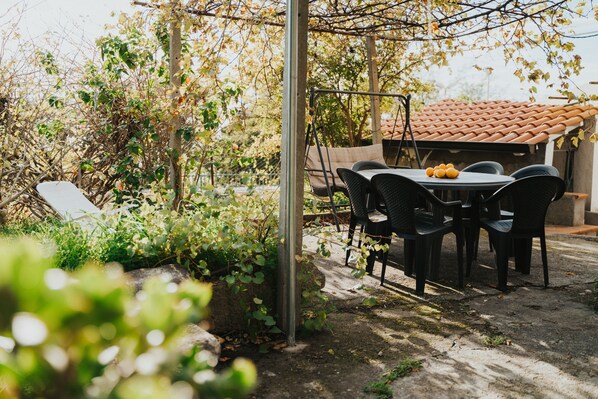 Un patio al aire libre con una mesa y sillas, rodeado de follaje y una pérgola.