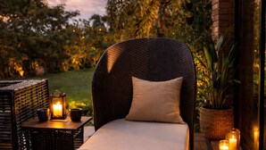 Una silla de mimbre con cojín, una mesita con un farol y velas en un patio con vistas a árboles y un edificio.