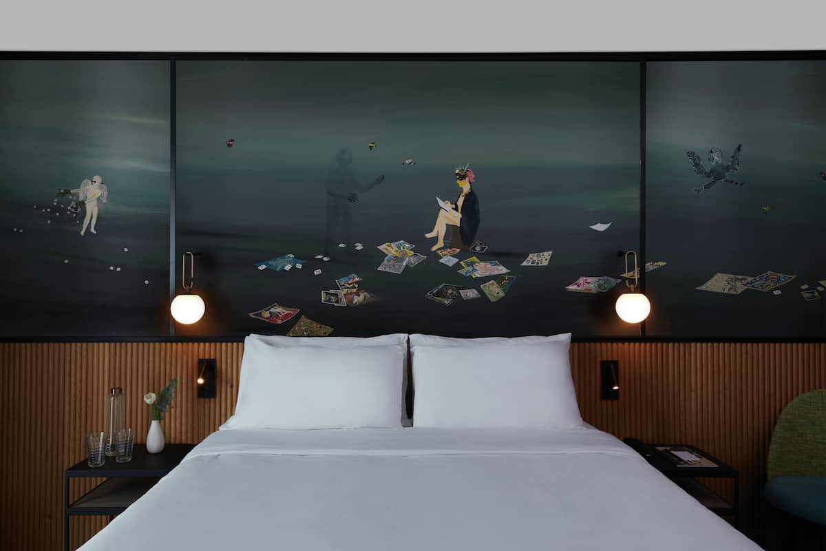 Habitación de hotel con un cuadro grande en la pared que muestra una escena surrealista, una cama bien hecha, dos mesitas de noche con lámparas y un panel de pared de madera.