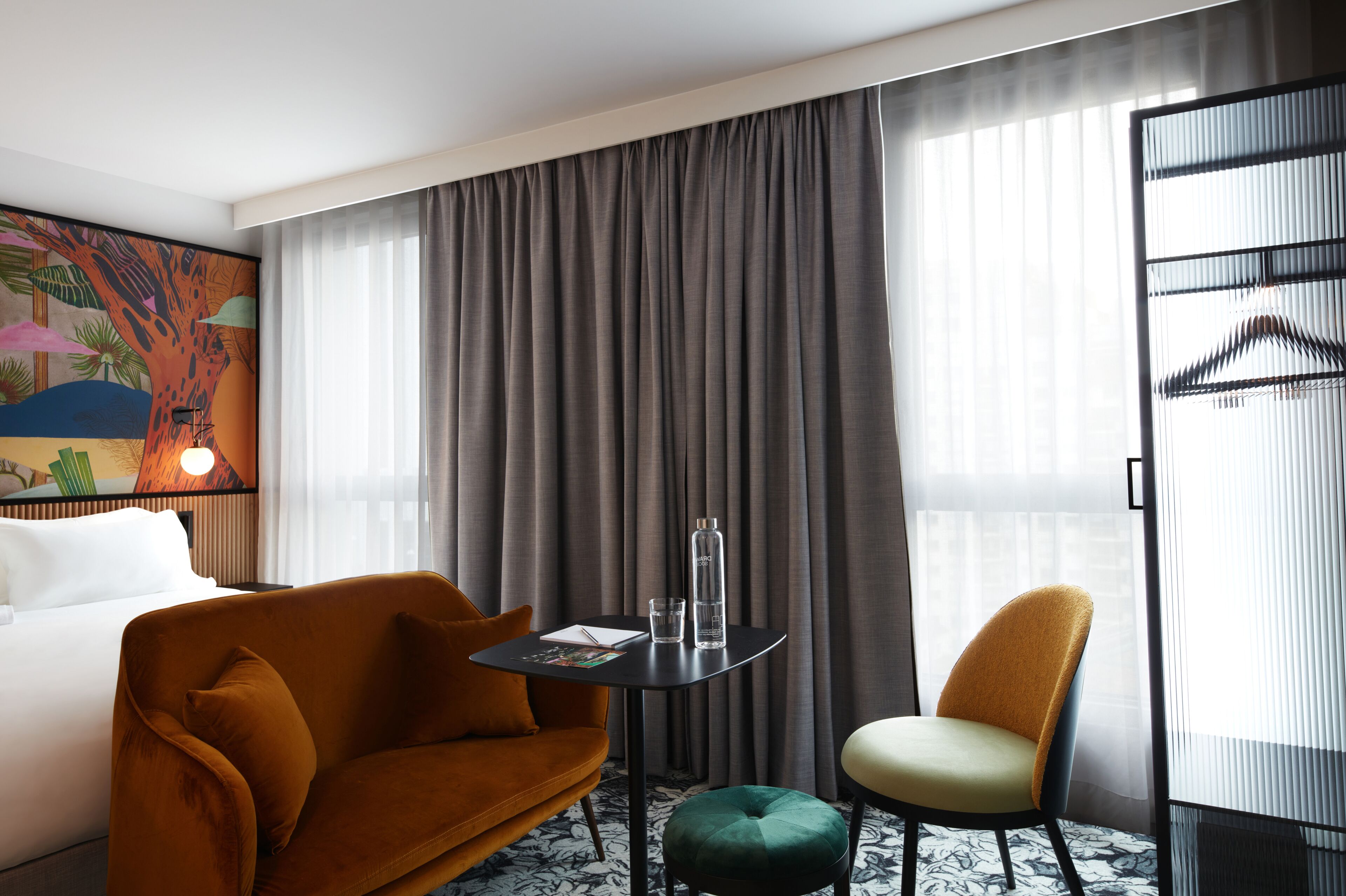 A hotel room with a large bed, a sofa, a chair, a small table with a bottle and glass, and a painting on the wall.
