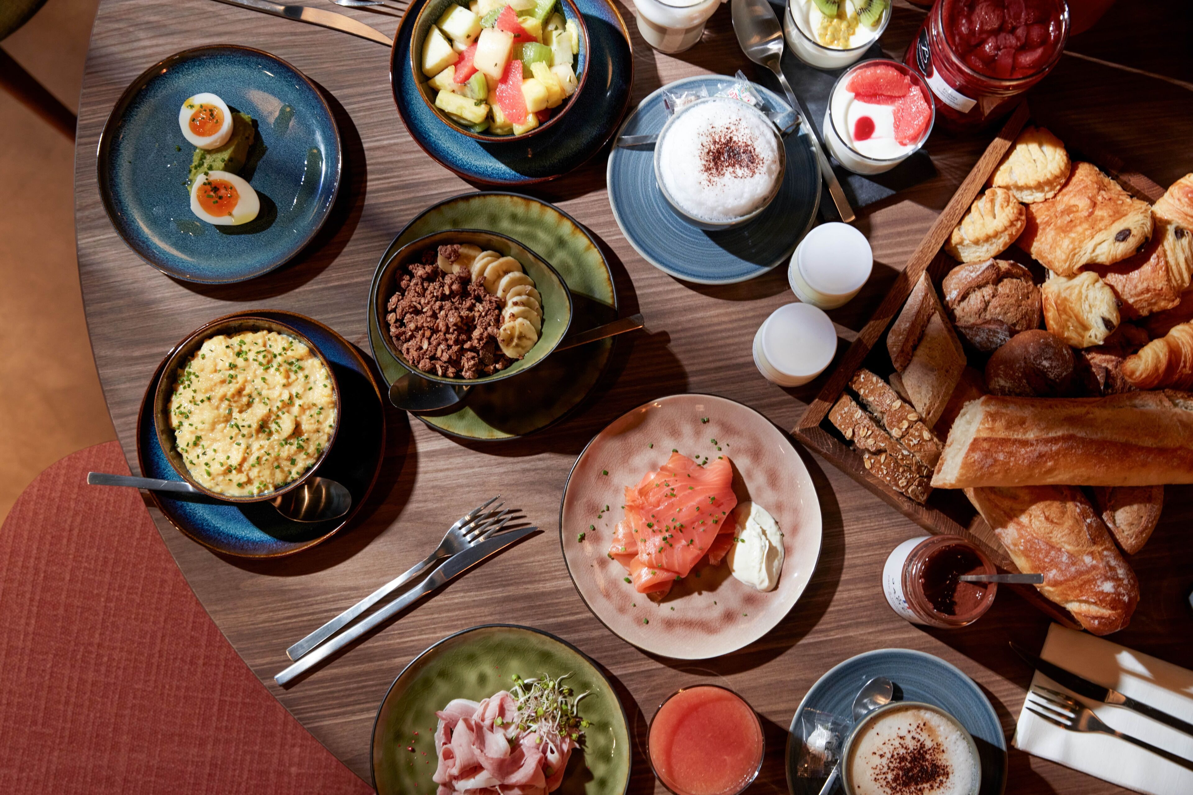 Diversos pratos de pequeno-almoço, incluindo ovos, salmão, pastelaria e fruta, sobre uma mesa de madeira.