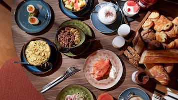 Diversos pratos de pequeno-almoço, incluindo ovos, salmão, pastelaria e fruta, sobre uma mesa de madeira.