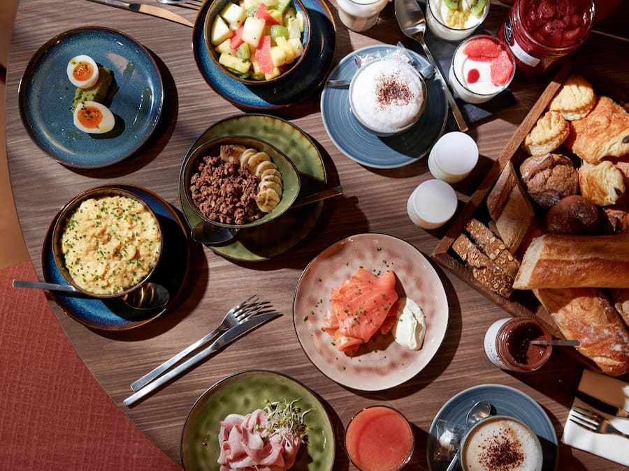 Diversos pratos de pequeno-almoço, incluindo ovos, salmão, pastelaria e fruta, sobre uma mesa de madeira.