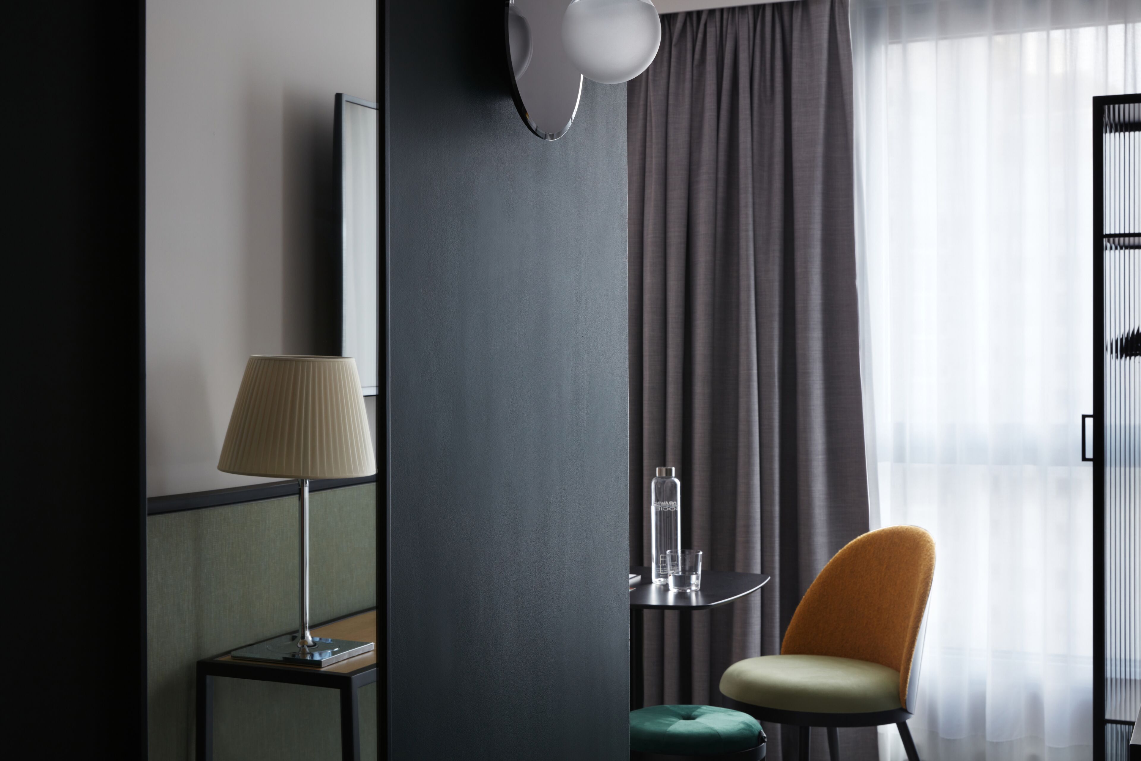 Camera d'hotel moderna con un letto, un comodino con una lampada, una sedia, un tavolino con una bottiglia e un bicchiere, e vista esterna filtrata da tende leggere.