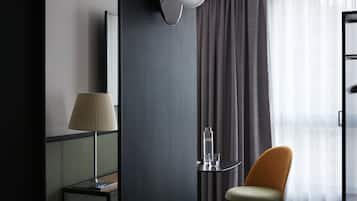 Camera d'hotel moderna con un letto, un comodino con una lampada, una sedia, un tavolino con una bottiglia e un bicchiere, e vista esterna filtrata da tende leggere.