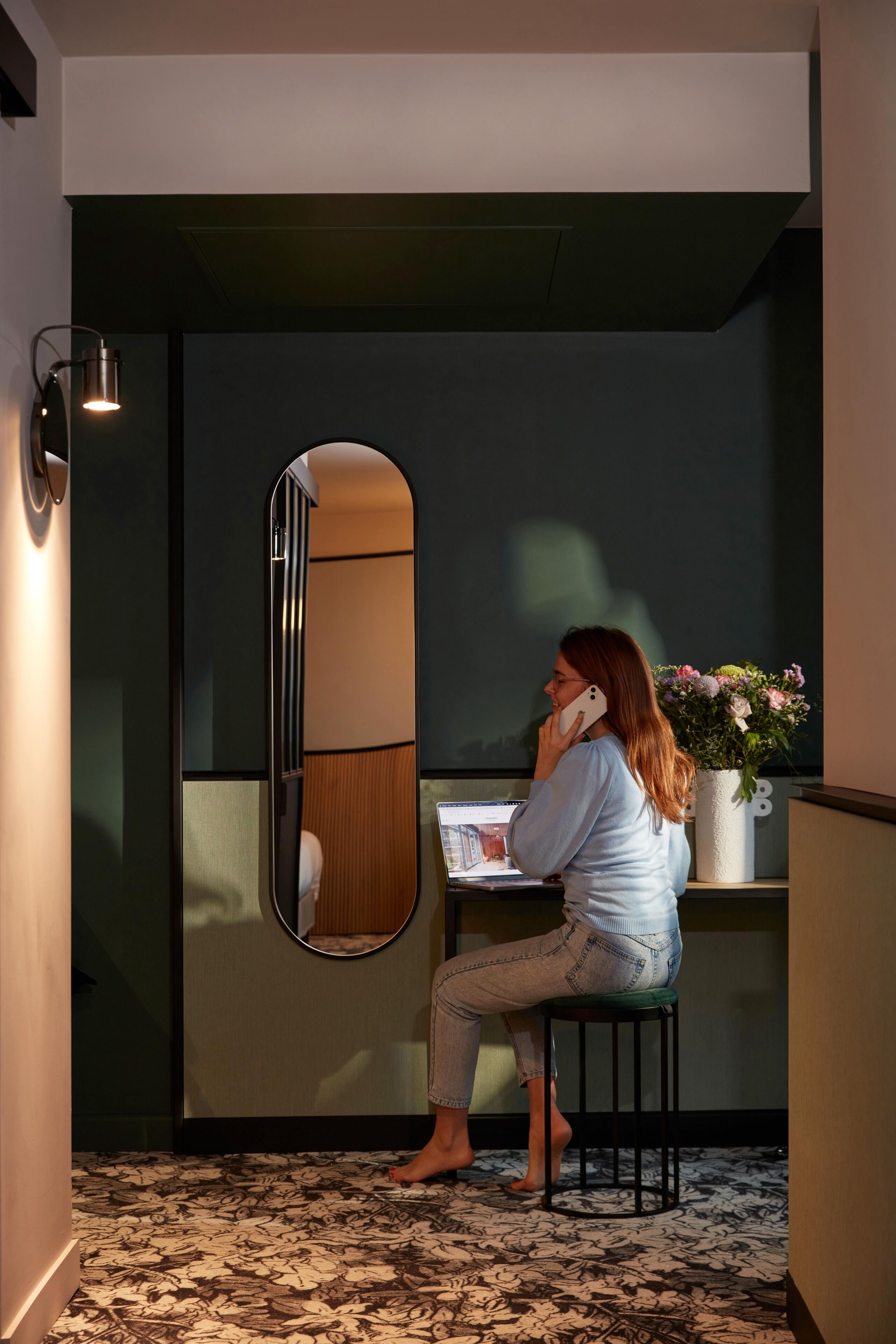 A woman sitting on a stool at a desk with a laptop, a mirror, and a vase of flowers.