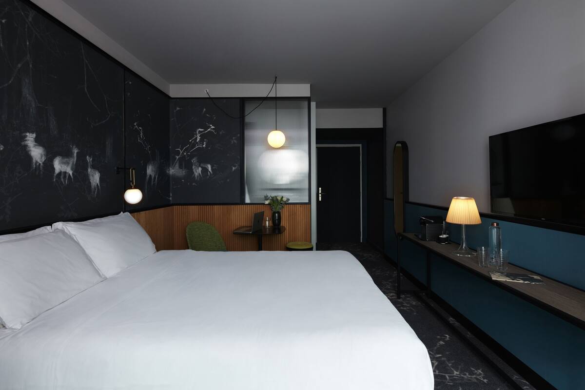 Una habitación de hotel moderna con una cama grande, un escritorio con lámpara, una mesita con una planta y un espejo amplio.