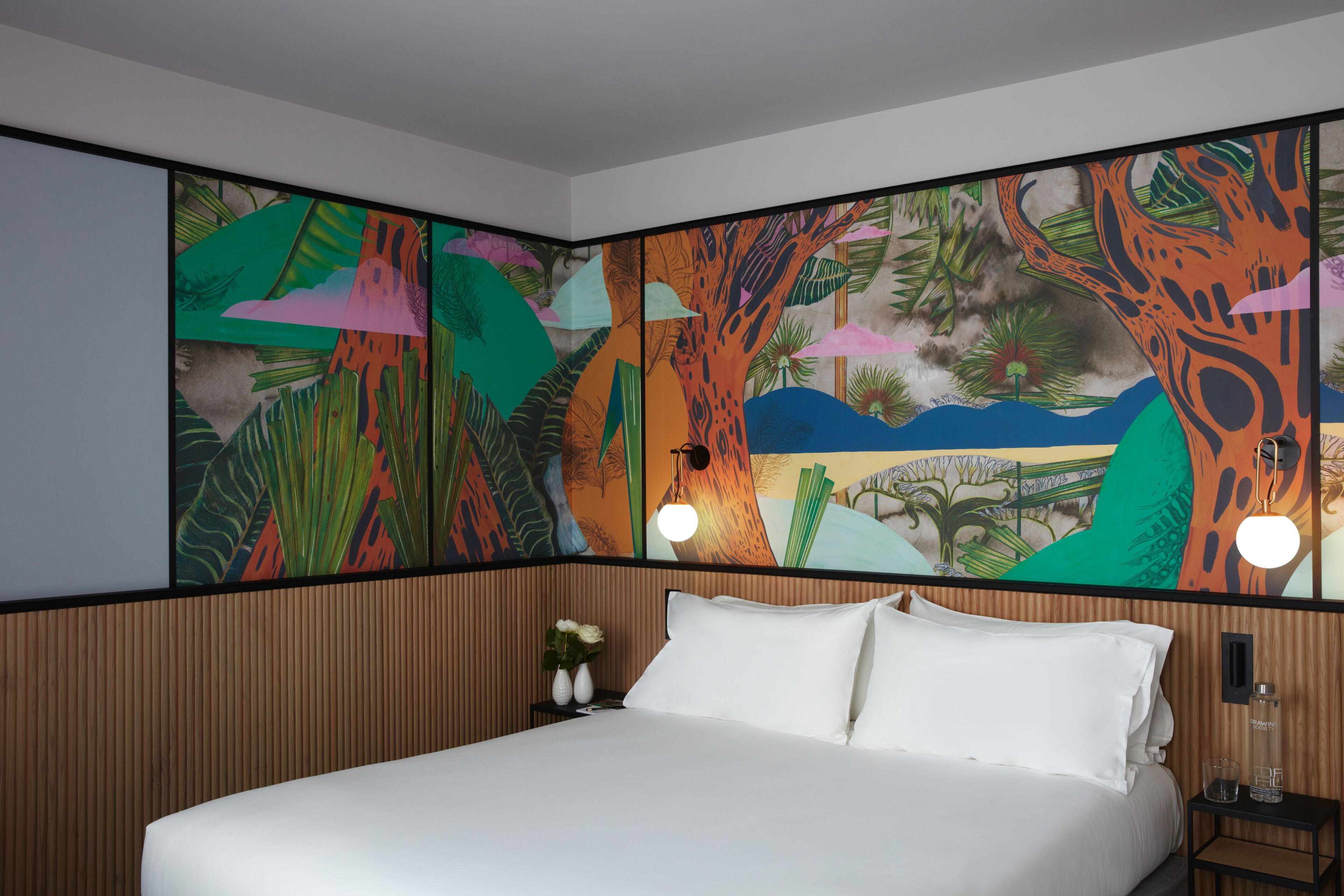 Una habitación de hotel con una cama grande, cabecera de madera, mesita de noche con un florero y un mural colorido en la pared.