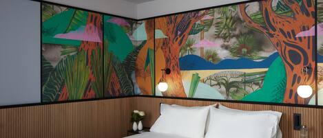 Una habitación de hotel con una cama grande, cabecera de madera, mesita de noche con un florero y un mural colorido en la pared.