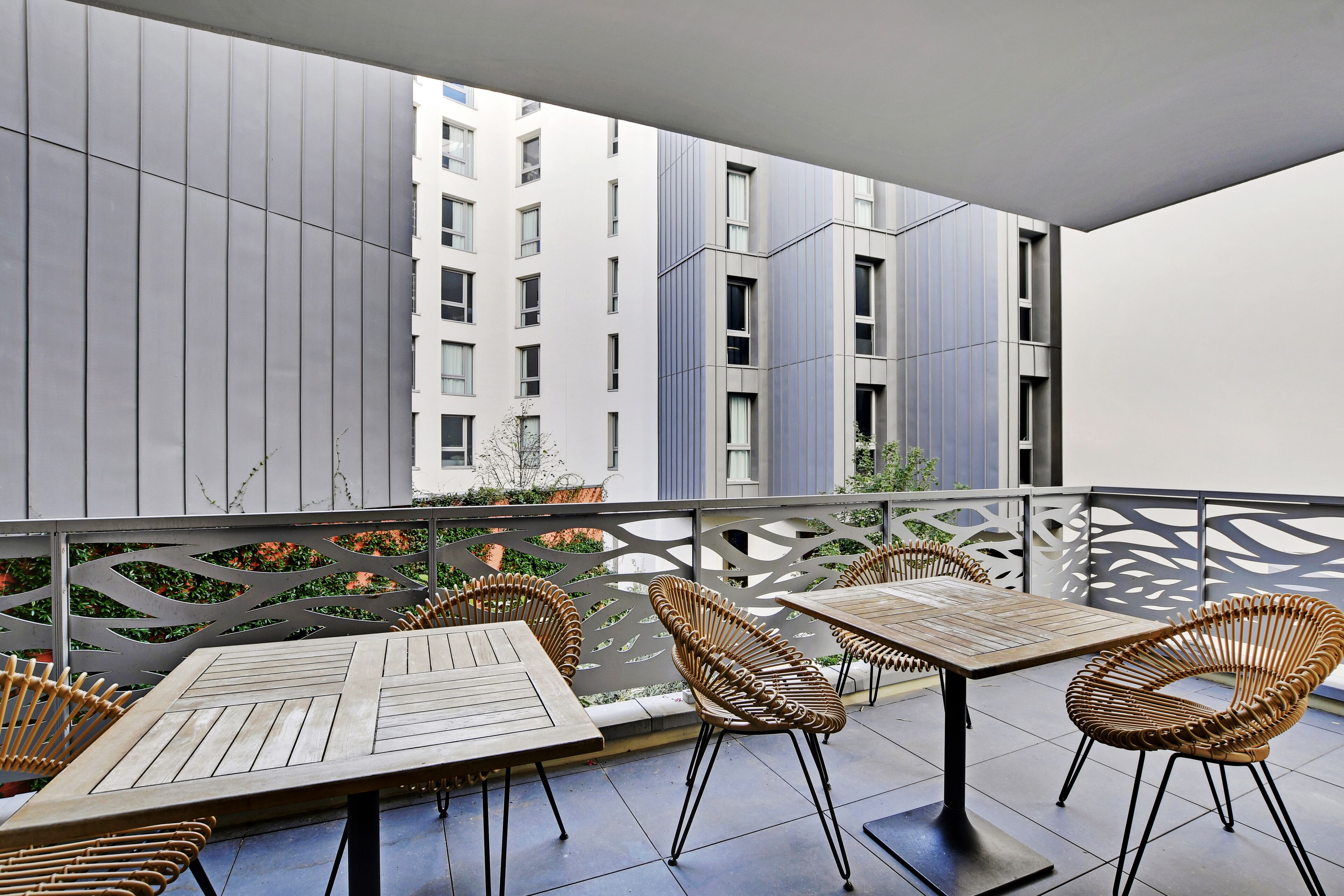 A balcony with outdoor seating, including tables and wicker chairs, overlooking a modern building with vertical siding and windows.