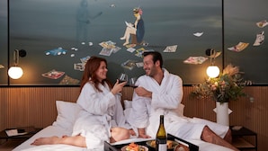 A couple in white robes sitting on a bed with a tray of food and wine, a vase of flowers, and a wall with cards and a painting. - Drawing House (Paris)