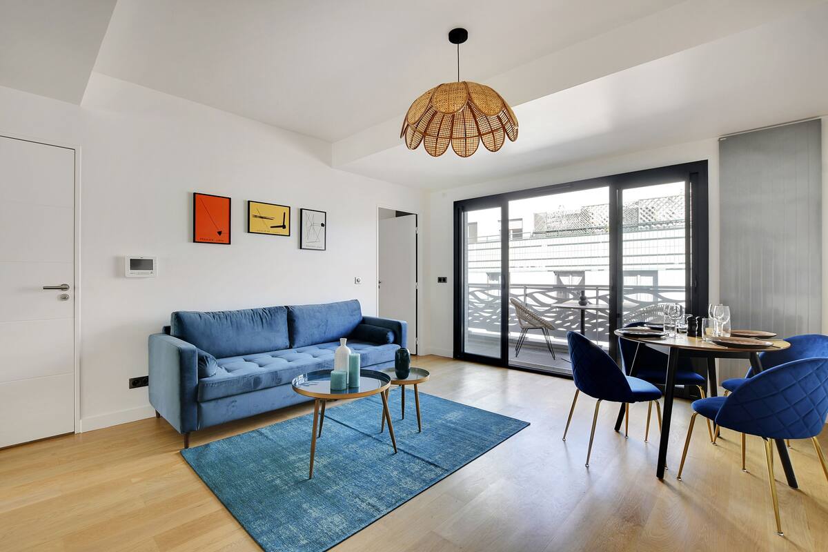 Una sala de estar moderna con un sofá azul, una mesa de centro y sillas azules. Hay un ventanal con vistas a edificios.