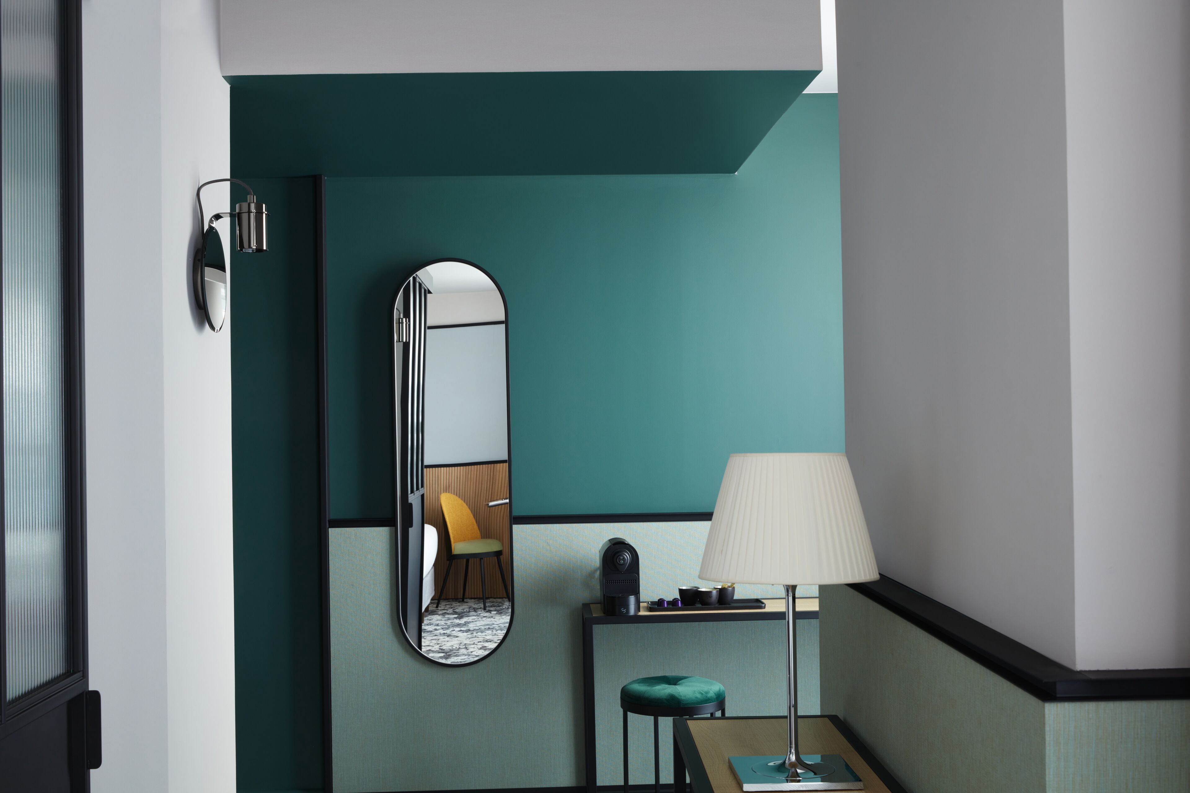 A hotel room with teal walls, a mirror, a lamp, a small table with a cushion, and a chair.