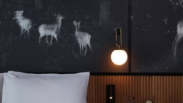 A hotel room with a bed, a nightstand with glasses and a vase, and a wall with a light fixture and a mural of deer.