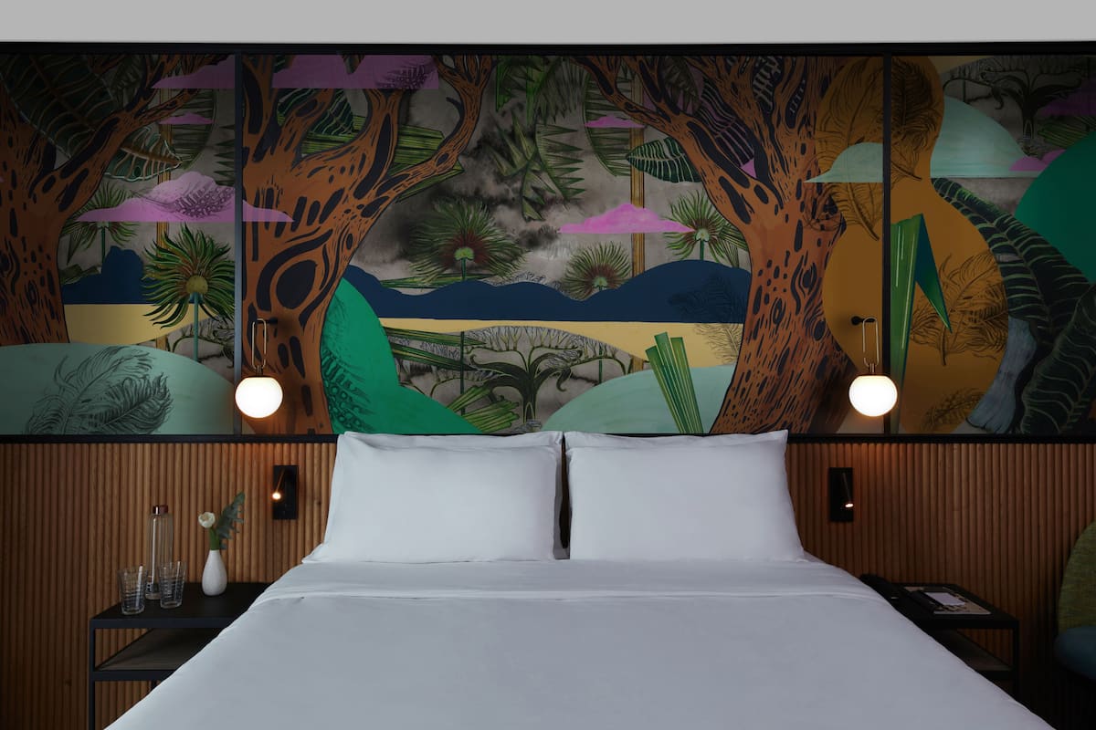 Una cama con sábanas blancas, una mesita de noche con lámpara y un mural en la pared que muestra una escena selvática.