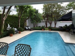 Una piscina rectangular con superficie de agua azul transparente, rodeada por una terraza blanca. Hay sillas de descanso y una pequeña estatua a la izquierda.