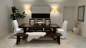 Een eetruimte met een donker houten tafel, witte stoelen en een bank. Aan weerszijden van de tafel hangen twee wandlampen.