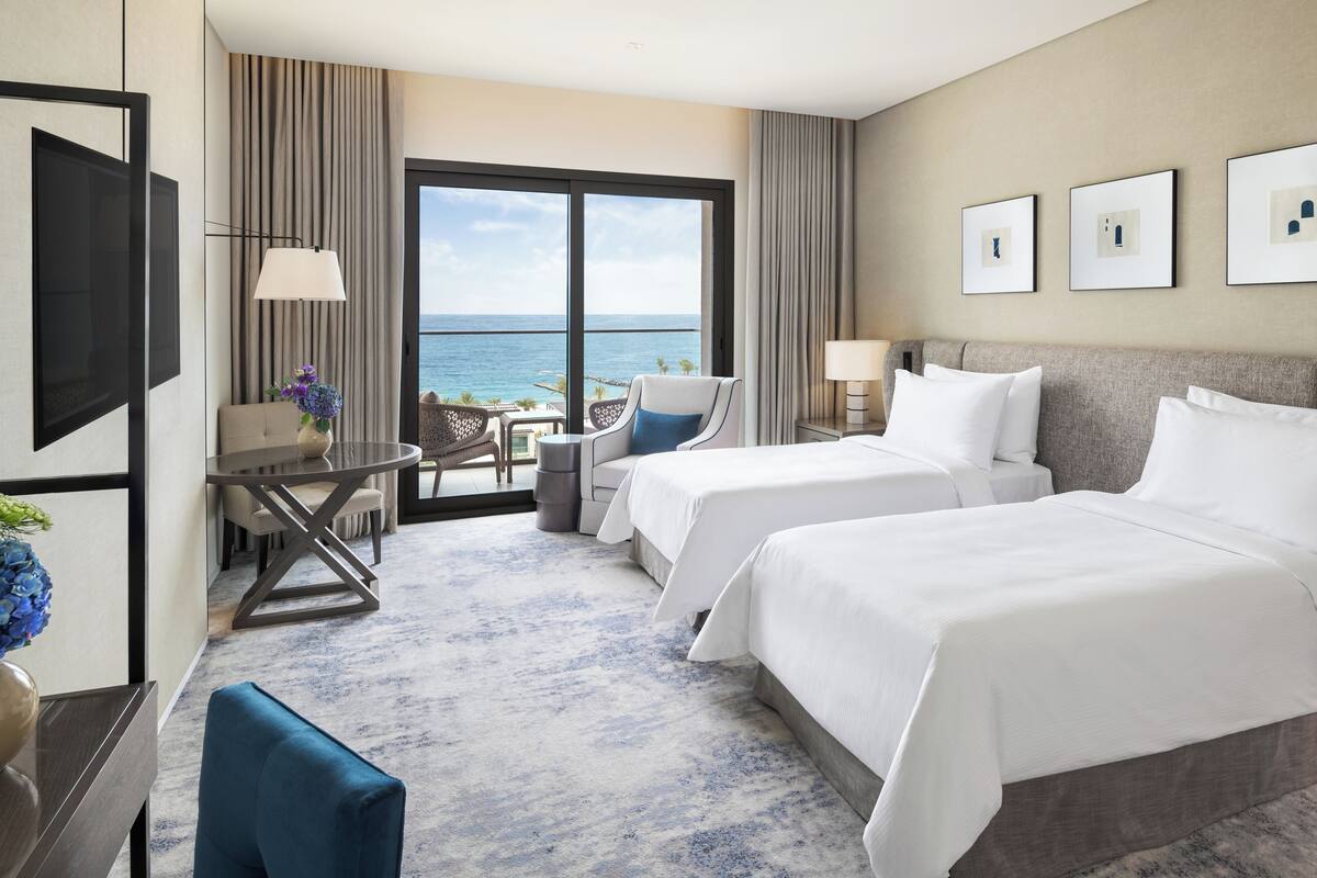 A hotel room with two beds, a desk, a chair, and a view of the ocean.