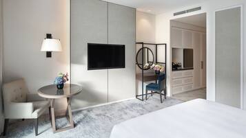 A hotel room with a bed, a round table with a lamp, a TV, a chair, and a small desk with a vase of flowers.