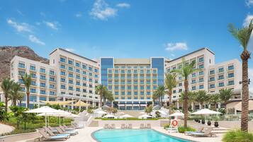 A hotel with a pool, sun loungers, umbrellas, and palm trees.