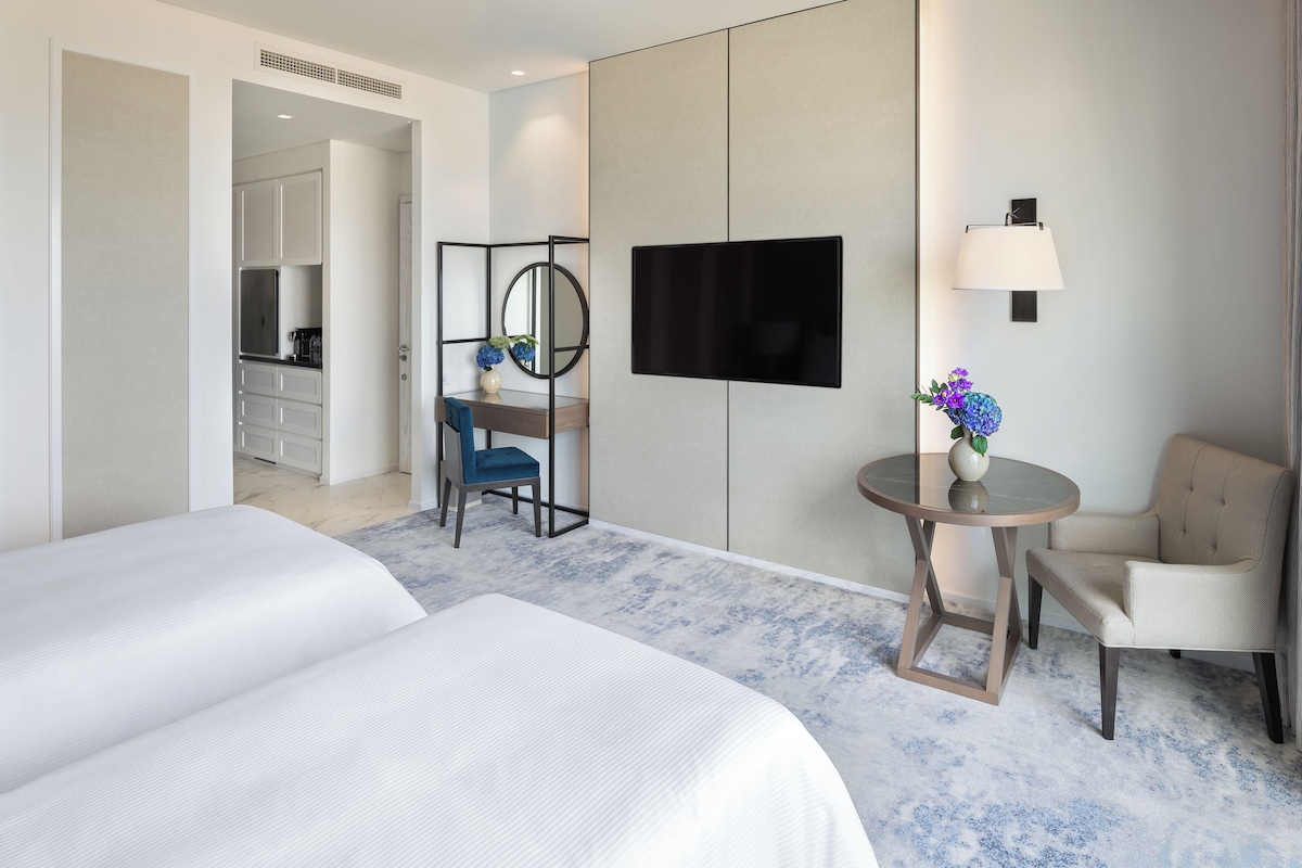 A hotel room with two beds, a desk with a chair, a round table with a chair, a TV mounted on the wall, and a small vase with flowers.