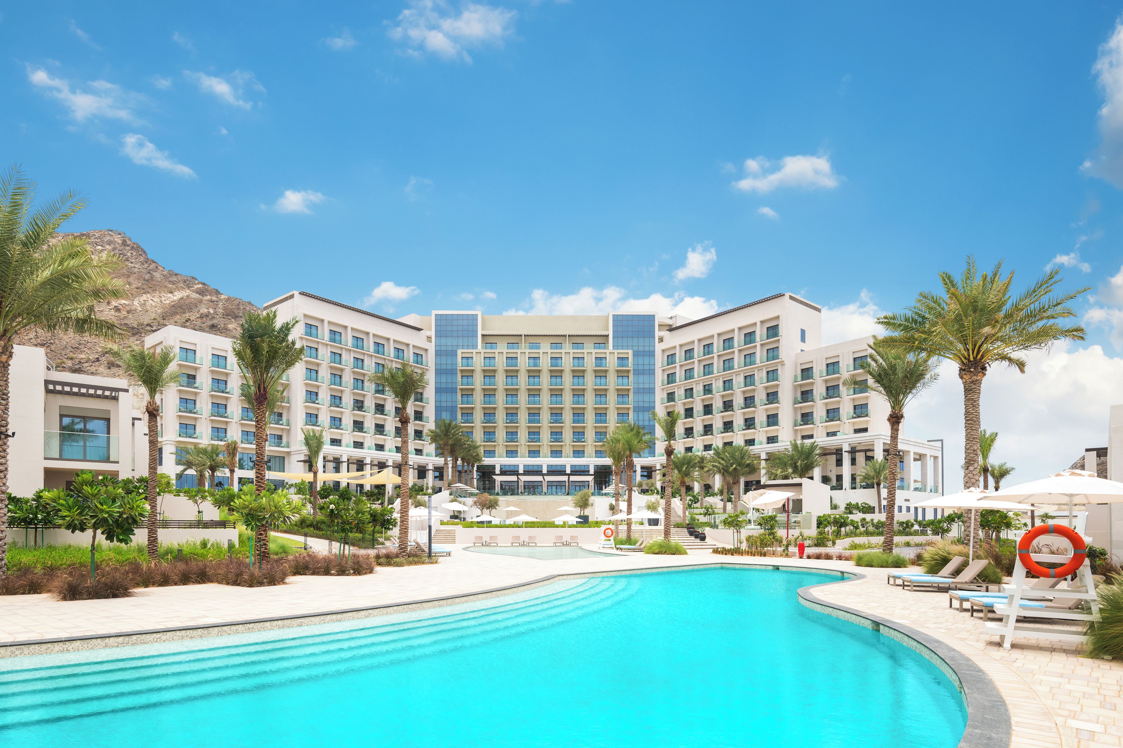 A hotel with a swimming pool, palm trees, and a mountainous backdrop.