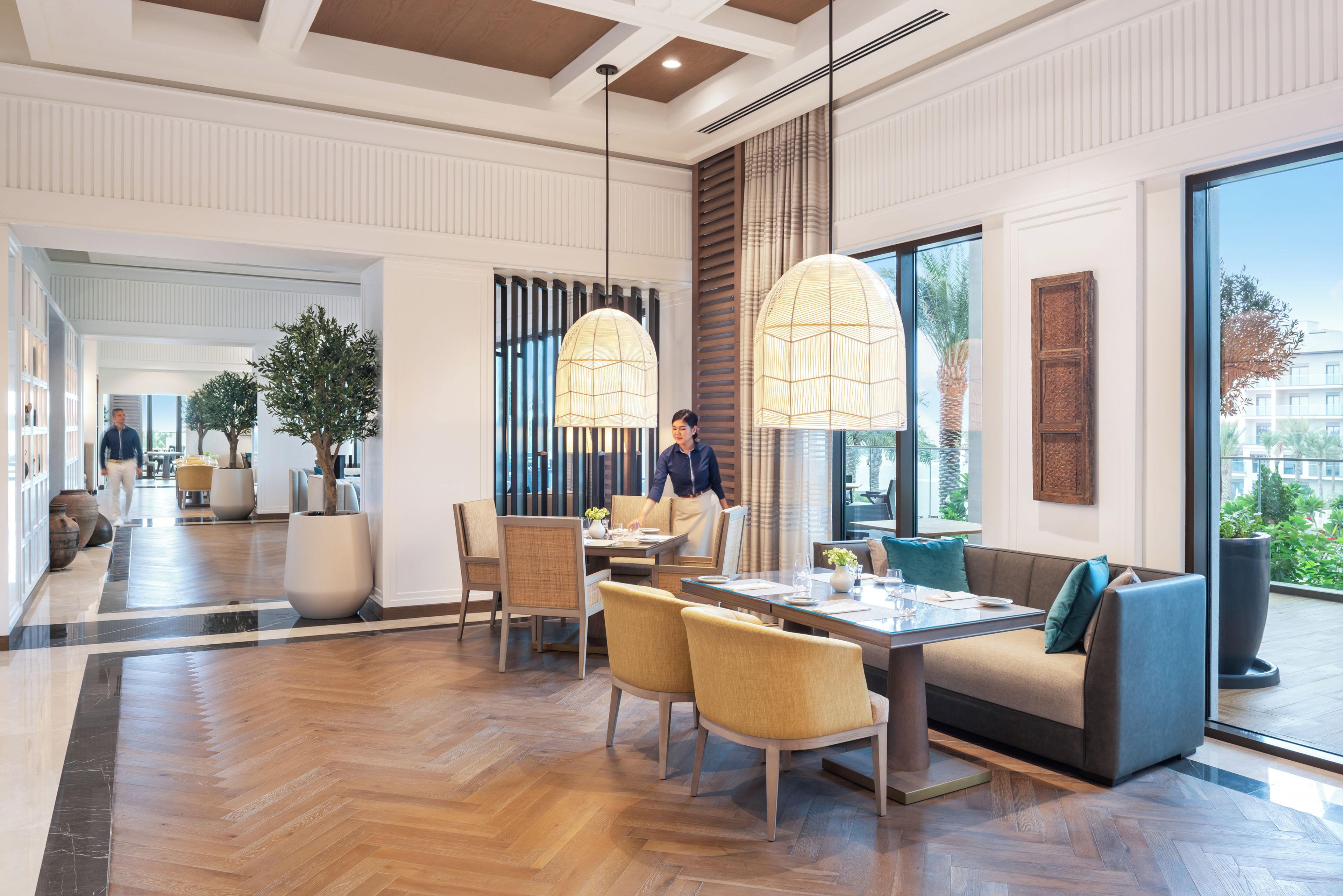A modern hotel lobby with a dining area, large windows, and a view of the outdoors.
