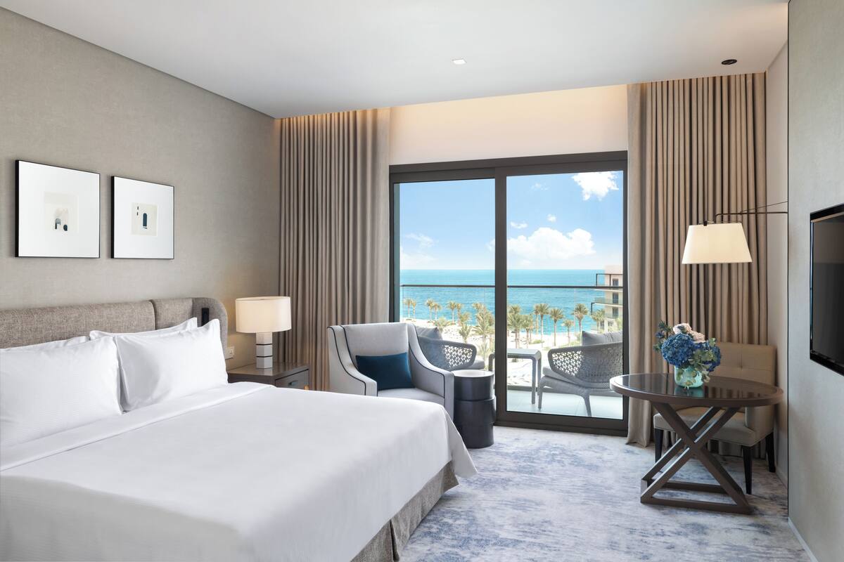 A hotel room with a large bed, a desk with a lamp, a chair, and a view of the ocean.