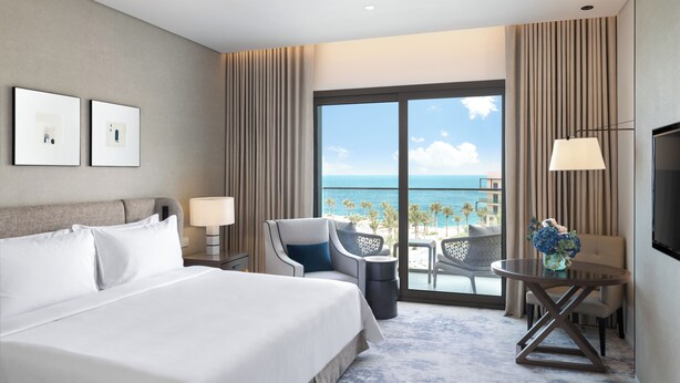 A hotel room with a large bed, a desk with a lamp, a chair, and a view of the ocean. - Address Beach Resort Fujairah (Al Aqah)