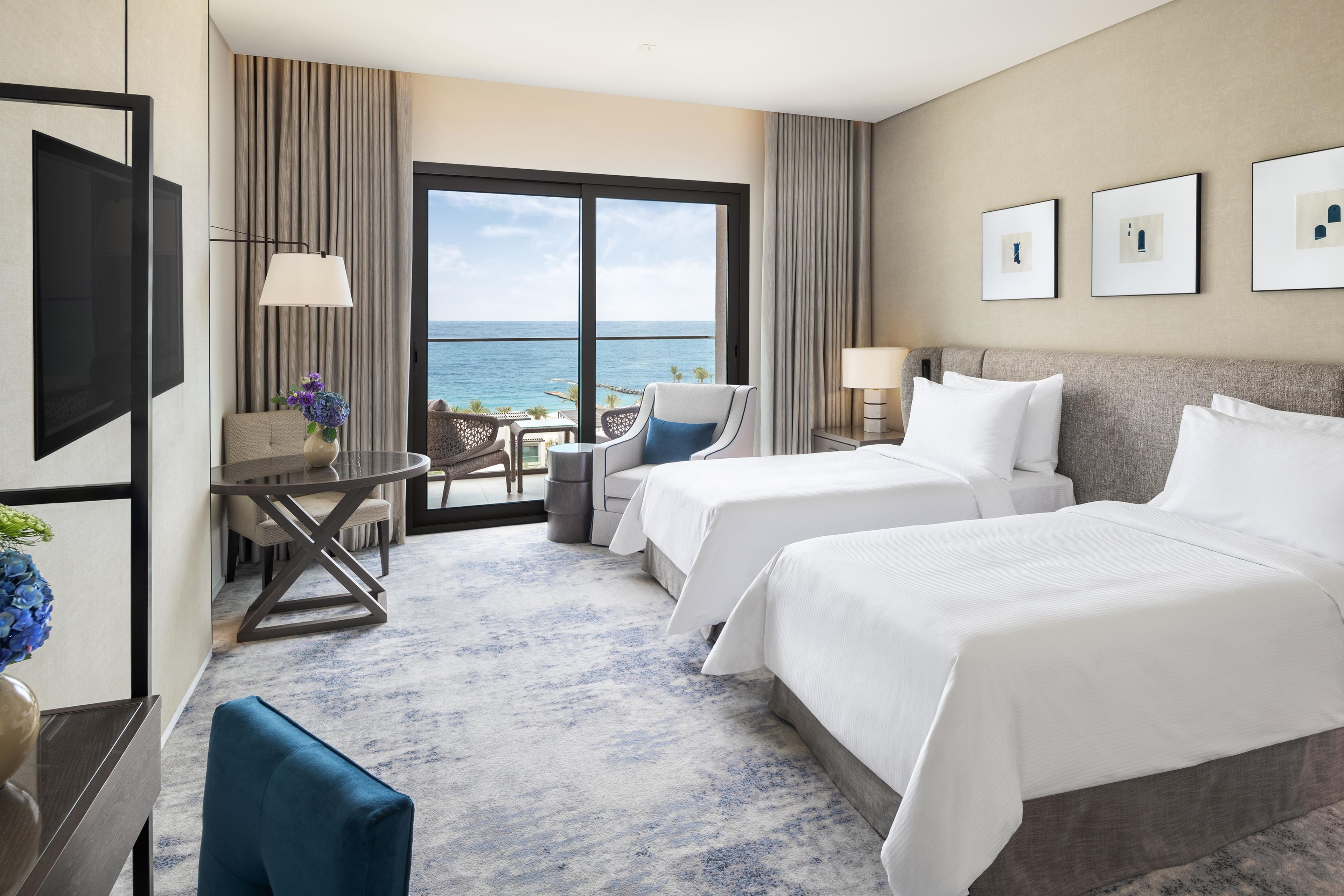 A hotel room with two beds, a desk, a chair, a view of the sea, and framed artwork on the wall.