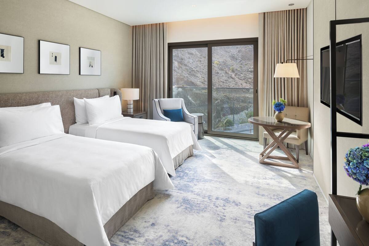 A hotel room with two beds, a chair, a small table, a TV, and a large window with a view.