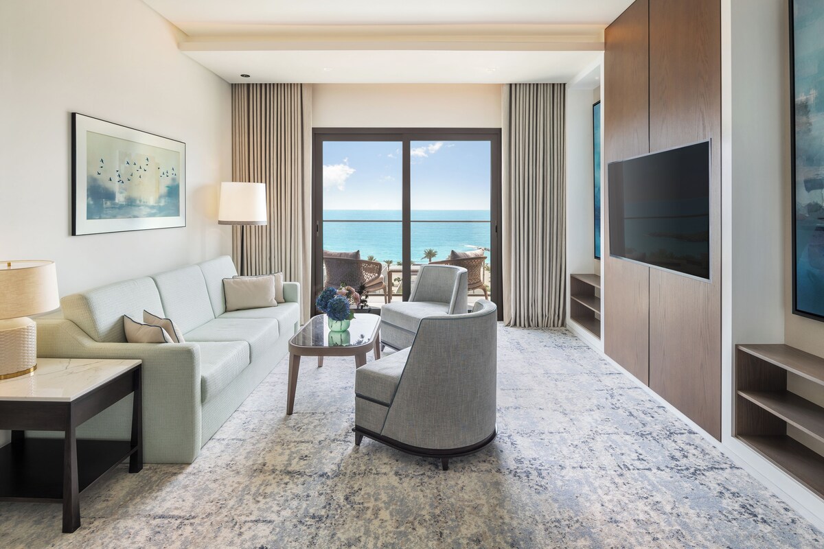 A modern living room with a sofa, armchair, and coffee table, a TV, and a view of the ocean.