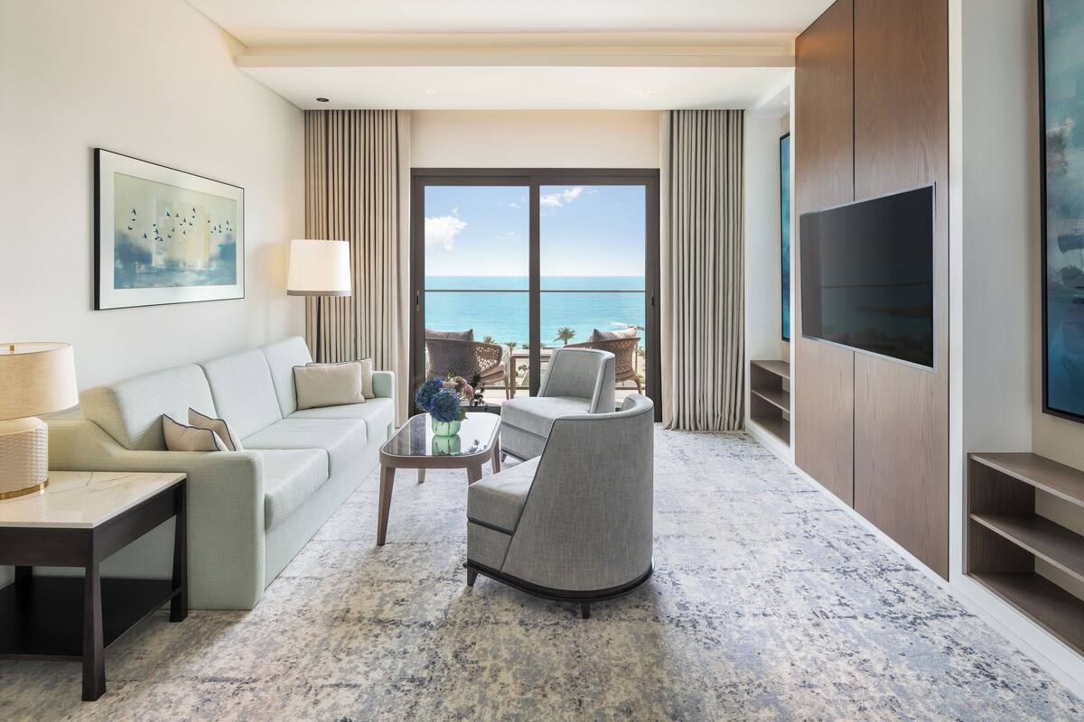 A modern living room with a sofa, armchair, and coffee table, a TV, and a view of the ocean.