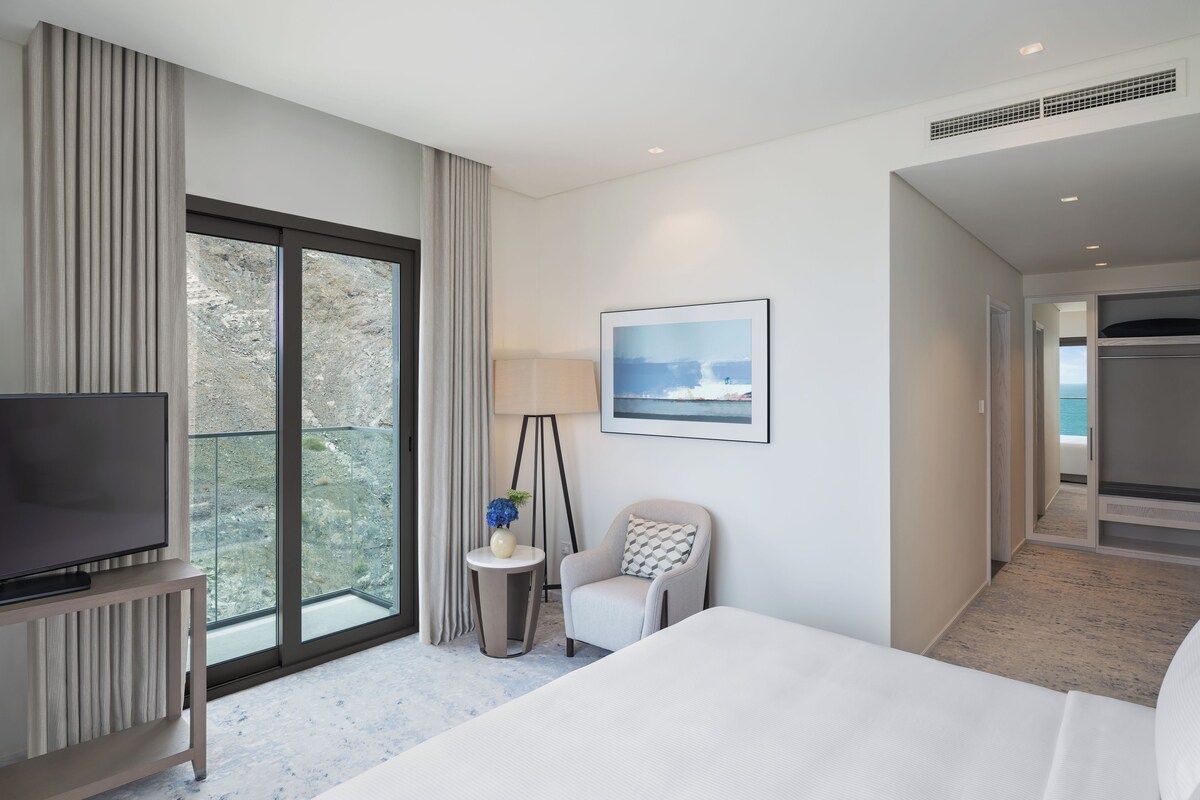 Une chambre d’hôtel avec un grand lit, un téléviseur posé sur un meuble, une petite table de chevet avec une lampe et un vase, une chaise, un tableau encadré au mur et une vue sur l’extérieur par une grande fenêtre.