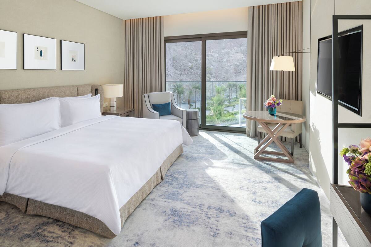 A hotel room with a large bed, a desk with a lamp, a TV, and a view of the outdoors.