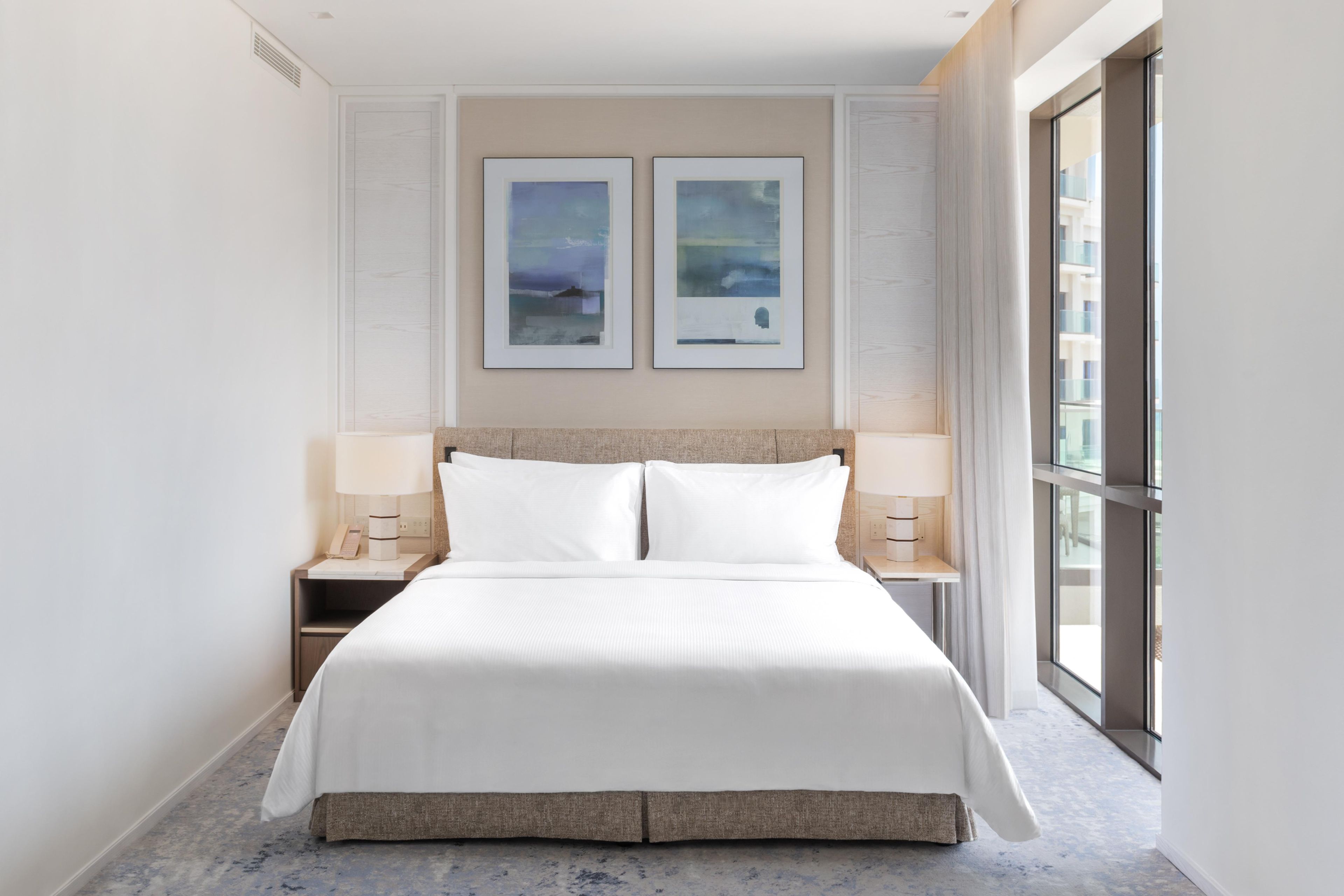 A hotel room with a large bed, two bedside tables, a nightstand, a window with a view, and two framed artworks on the wall.